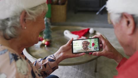 Smiling-senior-caucasian-couple-using-smartphone-for-christmas-video-call-with-man-on-screen