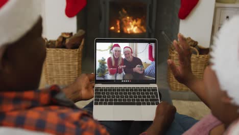 Afroamerikanisches-Paar-Mit-Weihnachtsmützen-Nutzt-Laptop-Für-Weihnachtsvideoanruf-Mit-Paar-Auf-Dem-Bildschirm