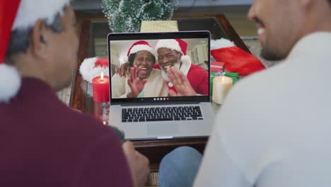 Smiling-biracial-father-and-son-using-laptop-for-christmas-video-call-with-happy-couple-on-screen
