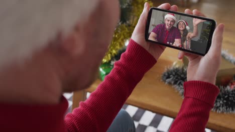 Kaukasischer-Mann-Mit-Weihnachtsmütze-Nutzt-Smartphone-Für-Weihnachtsvideoanruf-Mit-Lächelndem-Paar-Auf-Dem-Bildschirm
