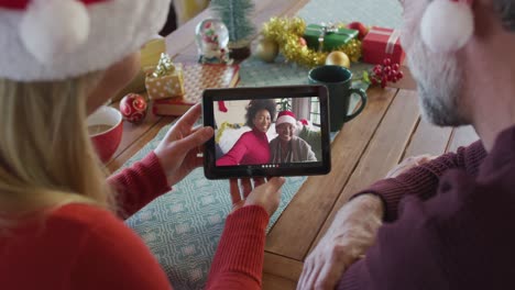 Smiling-caucasian-couple-with-santa-hats-using-tablet-for-christmas-video-call-with-family-on-screen