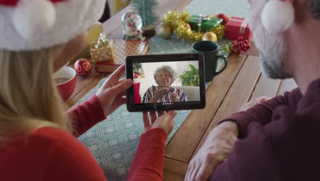 Lächelndes-Kaukasisches-Paar-Mit-Weihnachtsmützen,-Das-Ein-Tablet-Für-Einen-Weihnachtsvideoanruf-Mit-Einer-Frau-Auf-Dem-Bildschirm-Nutzt