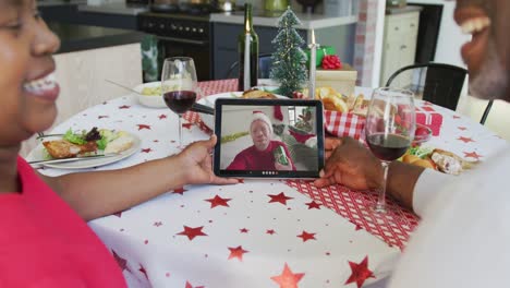African-american-couple-with-wine-using-tablet-for-christmas-video-call-with-happy-man-on-screen