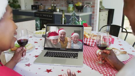 African-american-couple-with-wine-using-laptop-for-christmas-video-call-with-happy-family-on-screen