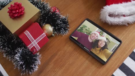 Smiling-diverse-couple-with-santa-hats-on-christmas-video-call-on-tablet