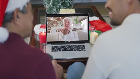 Smiling-biracial-father-with-son-using-laptop-for-christmas-video-call-with-senior-woman-on-screen