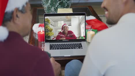Biracial-father-and-son-with-santa-hats-using-laptop-for-christmas-video-call-with-woman-on-screen