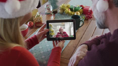 Lächelndes-Kaukasisches-Paar-Mit-Weihnachtsmützen,-Das-Ein-Tablet-Für-Einen-Weihnachtsvideoanruf-Mit-Einer-Frau-Auf-Dem-Bildschirm-Nutzt