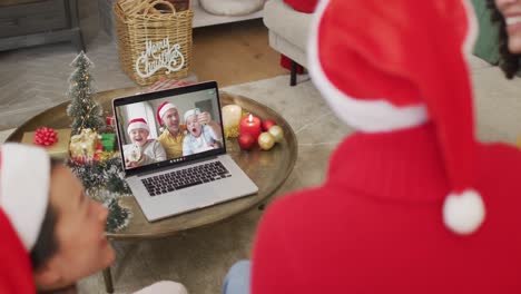 Vielfältige-Familie-Mit-Weihnachtsmützen-Nutzt-Laptop-Für-Weihnachtsvideoanruf-Mit-Lächelnder-Familie-Auf-Dem-Bildschirm