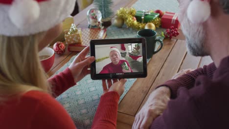 Lächelndes-Kaukasisches-Paar-Mit-Weihnachtsmützen,-Das-Ein-Tablet-Für-Einen-Weihnachtsvideoanruf-Mit-Einem-Mann-Auf-Dem-Bildschirm-Nutzt