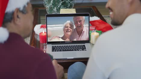 Biracial-father-and-son-with-santa-hats-using-laptop-for-christmas-video-call-with-couple-on-screen