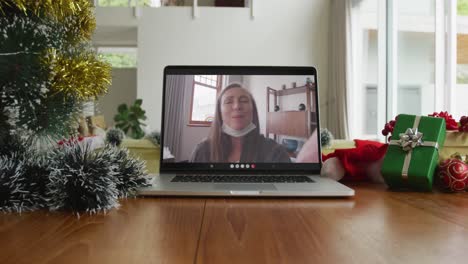 Smiling-caucasian-woman-with-face-mask-on-christmas-video-call-on-laptop