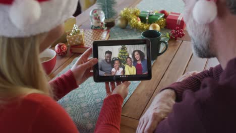 Smiling-caucasian-couple-with-santa-hats-using-tablet-for-christmas-video-call-with-family-on-screen