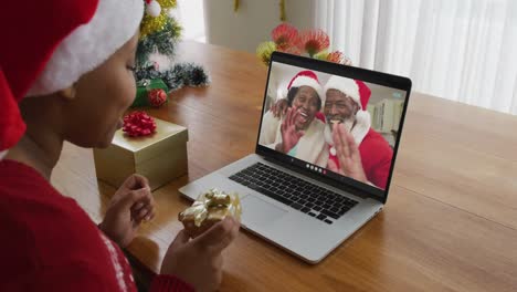 Afroamerikanische-Frau-Mit-Weihnachtsmütze-Nutzt-Laptop-Für-Weihnachtsvideoanruf-Mit-Paar-Auf-Dem-Bildschirm