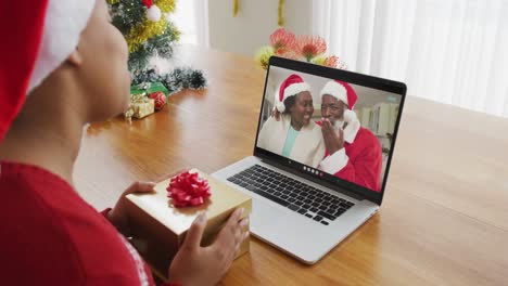 Mujer-Afroamericana-Con-Gorro-De-Papá-Noel-Usando-Una-Computadora-Portátil-Para-Una-Videollamada-Navideña,-Con-La-Familia-En-La-Pantalla