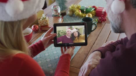 Kaukasisches-Paar-Mit-Weihnachtsmützen-Nutzt-Tablet-Für-Weihnachtsvideoanruf-Mit-Freunden-Auf-Dem-Bildschirm