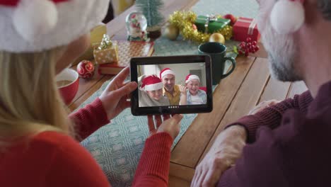 Lächelndes-Kaukasisches-Paar-Mit-Weihnachtsmützen,-Das-Ein-Tablet-Für-Einen-Weihnachtsvideoanruf-Mit-Der-Familie-Auf-Dem-Bildschirm-Nutzt