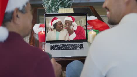 Smiling-biracial-father-and-son-using-laptop-for-christmas-video-call-with-couple-on-screen
