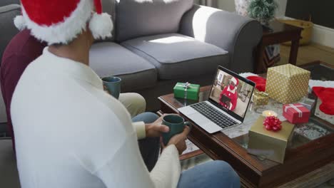 Biracial-father-and-son-with-santa-hats-using-laptop-for-christmas-video-call-with-santa-on-screen