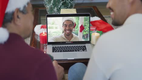 Smiling-biracial-father-and-son-using-laptop-for-christmas-video-call-with-man-on-screen