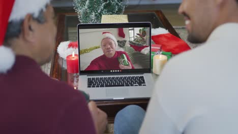 Smiling-biracial-father-with-son-using-laptop-for-christmas-video-call-with-man-on-screen