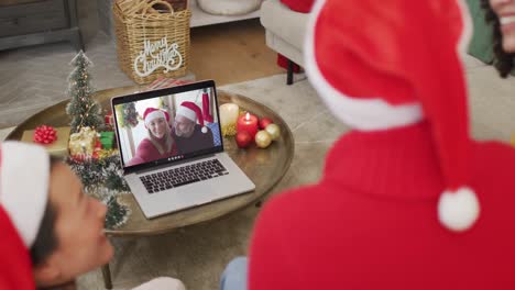 Vielfältige-Familie-Mit-Weihnachtsmützen-Nutzt-Laptop-Für-Weihnachtsvideoanruf-Mit-Glücklichem-Paar-Auf-Dem-Bildschirm