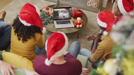 Diverse-family-with-santa-hats-using-laptop-for-christmas-video-call-with-boy-on-screen