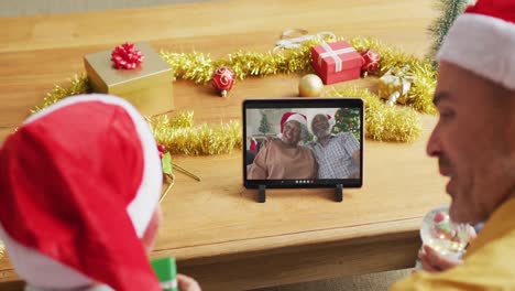 Kaukasischer-Vater-Und-Sohn-Mit-Weihnachtsmützen-Nutzen-Tablet-Für-Weihnachtsvideoanruf-Mit-Paar-Auf-Dem-Bildschirm