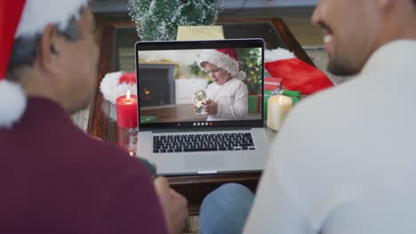 Biracial-father-and-son-with-santa-hats-using-laptop-for-christmas-video-call-with-boy-on-screen