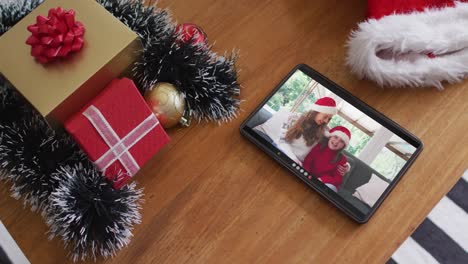 Smiling-caucasian-mother-and-daughter-wearing-santa-hats-on-christmas-video-call-on-tablet