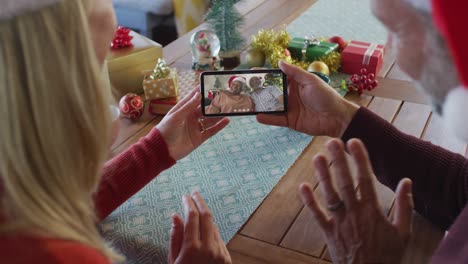 Kaukasisches-Paar-Mit-Weihnachtsmützen-Nutzt-Smartphone-Für-Weihnachtsvideoanruf-Mit-Paar-Auf-Dem-Bildschirm