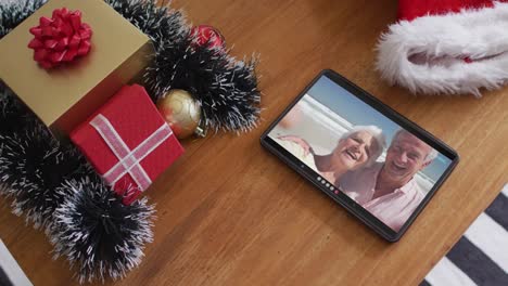 Smiling-senior-caucasian-couple-at-beach-on-christmas-video-call-on-tablet