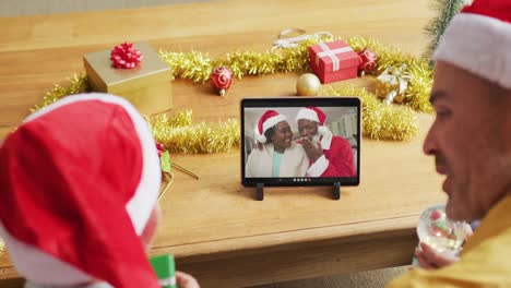 Kaukasischer-Vater-Und-Sohn-Mit-Weihnachtsmützen-Nutzen-Tablet-Für-Weihnachtsvideoanruf-Mit-Paar-Auf-Dem-Bildschirm
