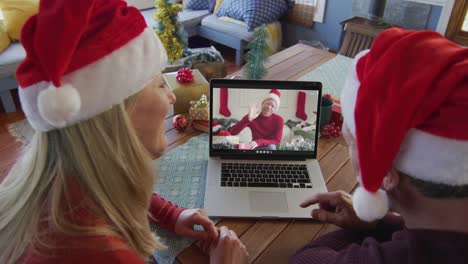 Smiling-caucasian-couple-with-santa-hats-using-laptop-for-christmas-video-call-with-man-on-screen