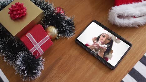 Smiling-caucasian-woman-wearing-santa-hat-and-blowing-snow-on-christmas-video-call-on-tablet
