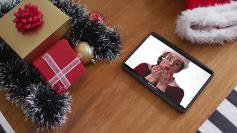 Caucasian-woman-wearing-santa-hat-and-blowing-snow-on-christmas-video-call-on-tablet