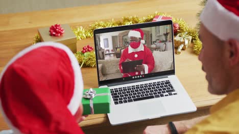 Caucasian-father-and-son-with-santa-hats-using-laptop-for-christmas-video-call-with-santa-on-screen