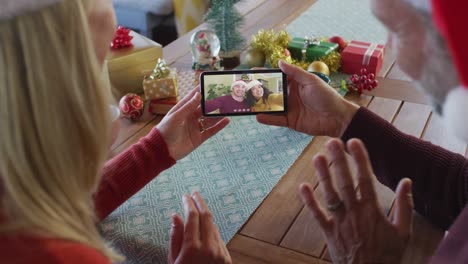 Kaukasisches-Paar-Mit-Weihnachtsmützen-Nutzt-Smartphone-Für-Weihnachtsvideoanruf-Mit-Paar-Auf-Dem-Bildschirm