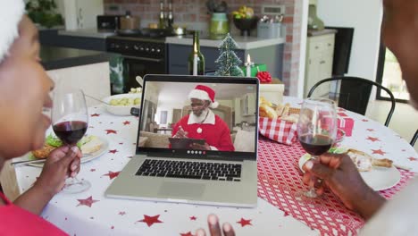 African-american-couple-with-wine-using-laptop-for-christmas-video-call-with-santa-on-screen