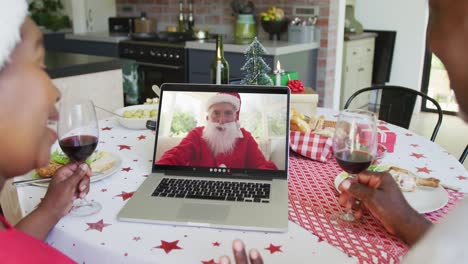African-american-couple-with-wine-using-laptop-for-christmas-video-call-with-happy-santa-on-screen