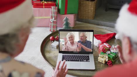 Senior-caucasian-couple-with-santa-hats-using-laptop-for-christmas-video-call-with-couple-on-screen