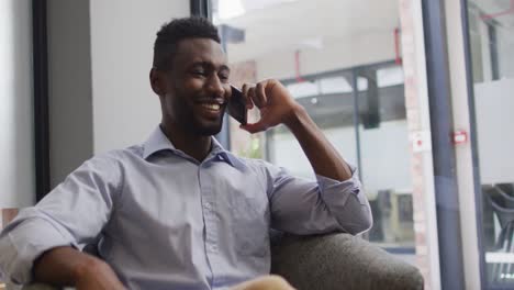Lächelnder-Afroamerikanischer-Geschäftsmann,-Der-Im-Sessel-Sitzt-Und-Im-Modernen-Büro-Auf-Dem-Smartphone-Spricht