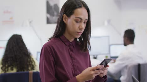Portrait-of-smiling-biracial-businesswoman-using-smartphone-looking-at-camera-in-modern-office