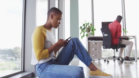 African-american-creative-businesswoman-sitting-on-floor-using-smartphone,-male-colleague-at-desk