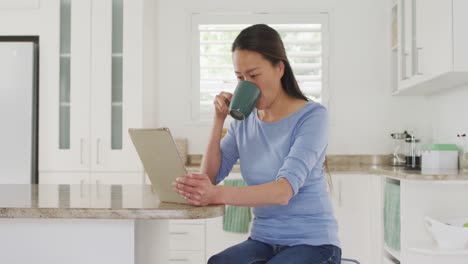 Happy-asian-woman-sitting-at-table,-drinking-coffee-and-using-tablet-in-kitchen