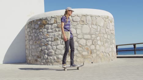 Video-Eines-Kaukasischen-Mannes-Mit-Dreadlocks,-Der-Auf-Der-Sonnigen-Strandpromenade-Skateboard-Fährt