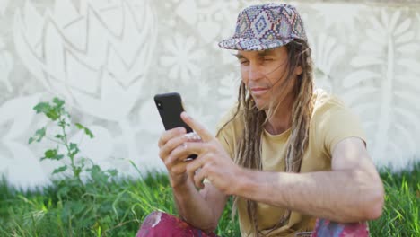 Video-Eines-Lächelnden-Kaukasischen-Mannes-Mit-Dreadlocks,-Der-Sein-Smartphone-Benutzt-Und-Im-Gras-Sitzt,-Neben-Einem-Wandgemälde