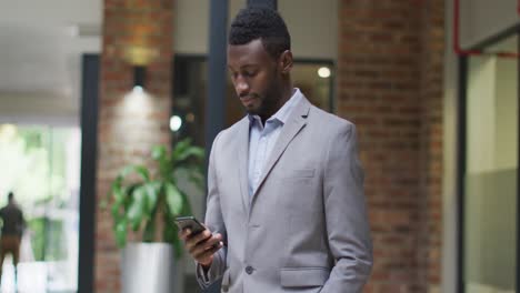 Smiling-african-american-businessman-standing-in-corridor-using-smartphone-in-modern-office