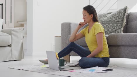 Mujer-Asiática-Enfocada-Sentada-En-El-Suelo-Y-Trabajando-Remotamente-Desde-Casa-Con-Teléfono-Inteligente-Y-Computadora-Portátil