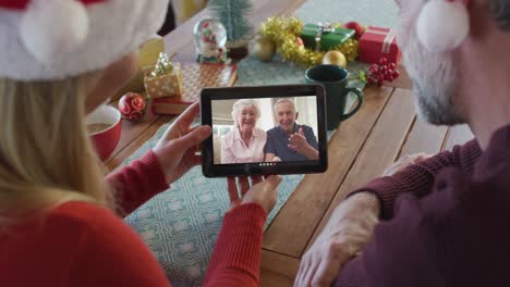 Caucasian-couple-with-santa-hats-using-tablet-for-christmas-video-call-with-couple-on-screen